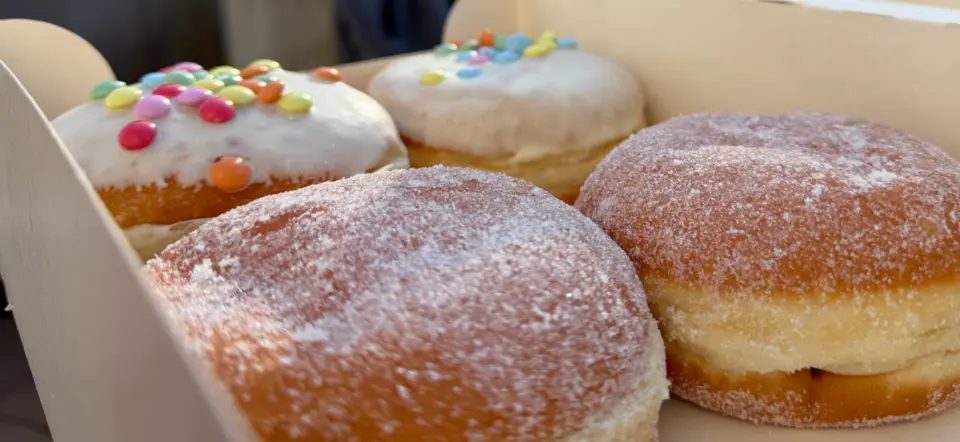 Foto eines geöffneten Kartons mit vier Donuts (oder Berlinern), reich verziert mit Zuckerguss und Schokolinsen. Das Bild visualisiert die "Zucker-Seite" im Blog-Vergleich zwischen der oft freiwilligen Lebensmittelkennzeichnung (wie Nutri-Score) und der strengen Regulierung von unsichtbaren Klima-Claims.