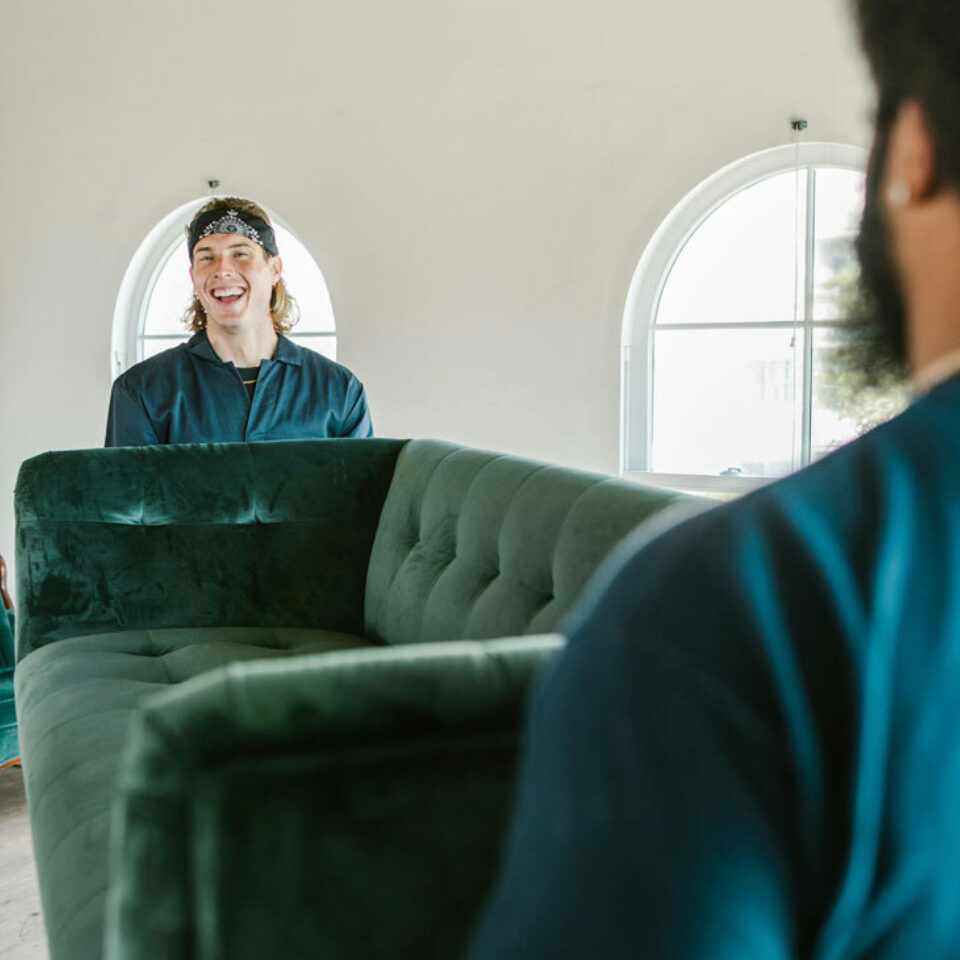 Zwei Männer tragen gemeinsam ein grünes Sofa durch ein helles Zimmer mit Umzugskartons.