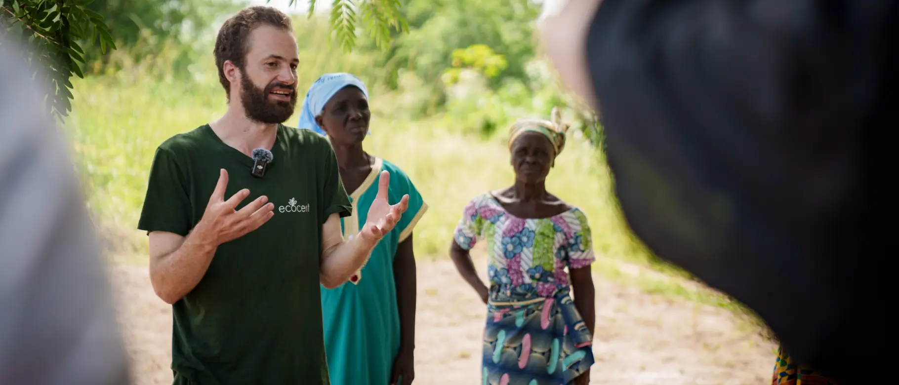 Foto des natureOffice Projektleiters vor Ort in Togo, der Besuchern gestenreich die sozialen und ökologischen Maßnahmen des Klimaschutzprojekts erklärt. Im Hintergrund sind zwei Frauen aus der lokalen Gemeinschaft zu sehen – ein Beleg für transparente Arbeit und Verankerung vor Ort.
