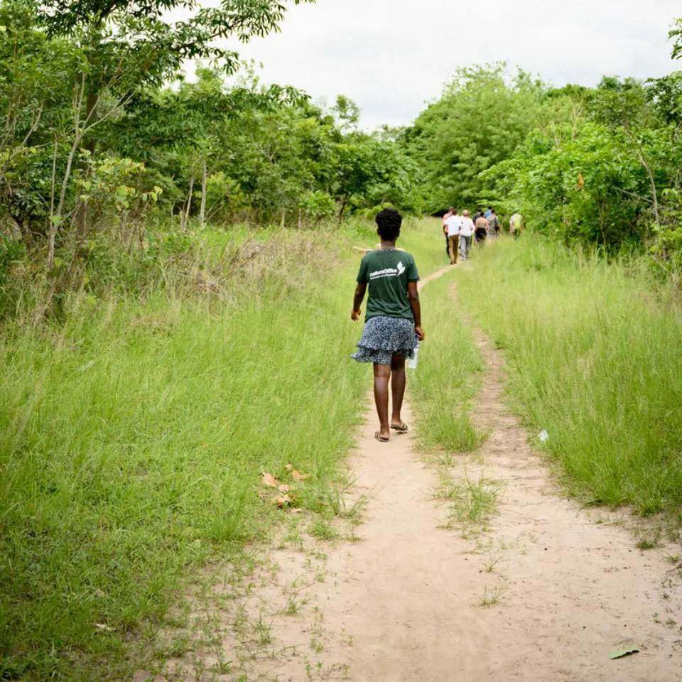Mädchen geht barfuß einen sandigen Weg entlang, Gruppe Menschen vorne, grüne Vegetation.