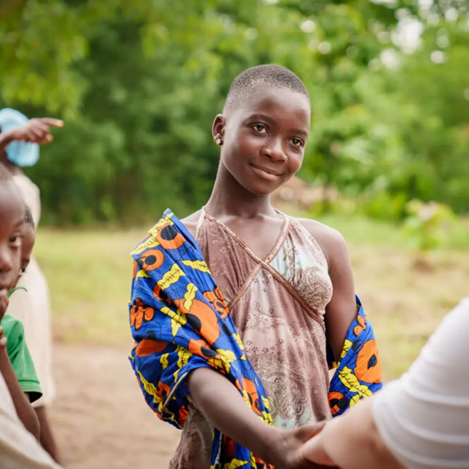 Mädchen in afrikanischer Kleidung schüttelt draußen einer Frau die Hand.