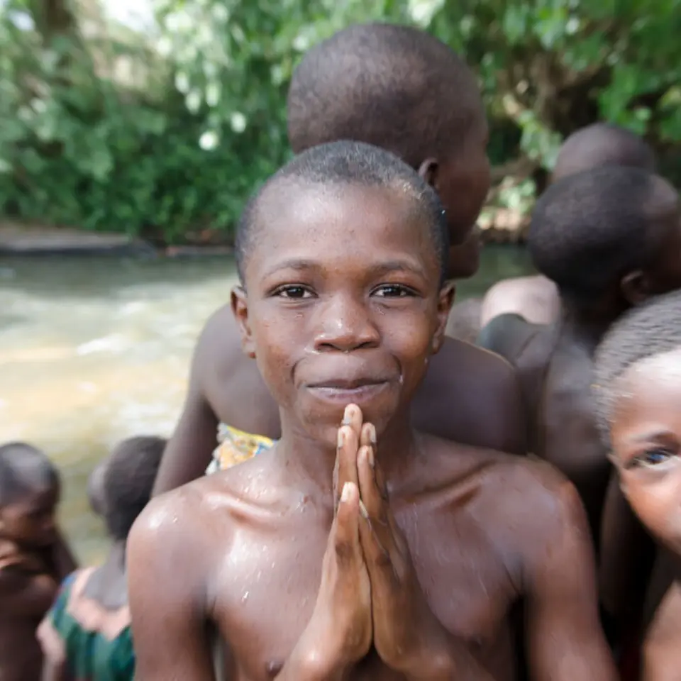 Junge mit gefalteten Händen steht lachend in einer Gruppe Kinder am Fluss.