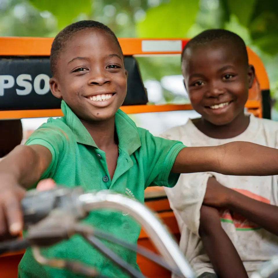 Zwei lachende Kinder auf einem Tricycle im PROJECT TOGO: Ein Beispiel für die soziale Unterstützung (Contribution) durch Partner wie Gebrüder Weiss im Klimaschutzprojekt in Togo.