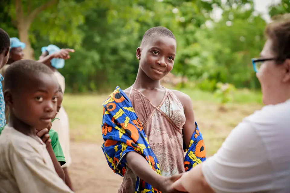 Mädchen in afrikanischer Kleidung schüttelt draußen einer Frau die Hand.