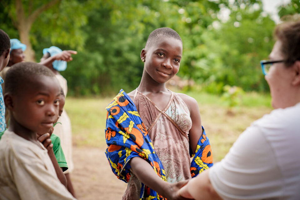 Mädchen in afrikanischer Kleidung schüttelt draußen einer Frau die Hand.