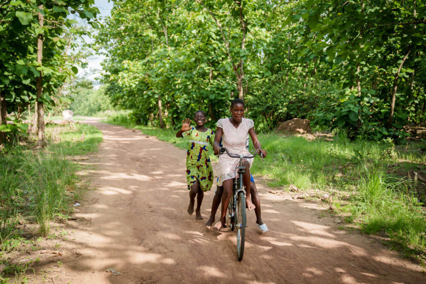 Mädchen fährt Fahrrad auf Sandweg, weiteres Mädchen läuft lachend daneben.