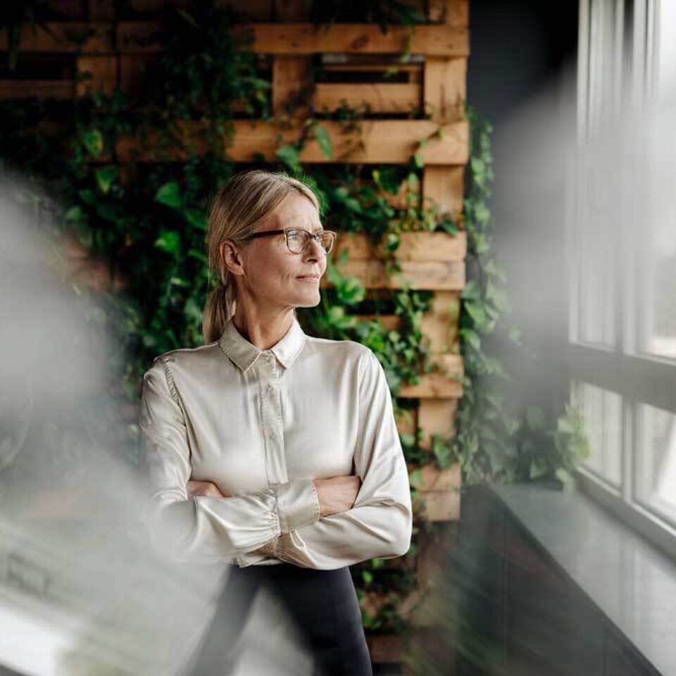 Frau mit Brille blickt nachdenklich aus einem Fenster, im Hintergrund grüne Pflanzenwand.