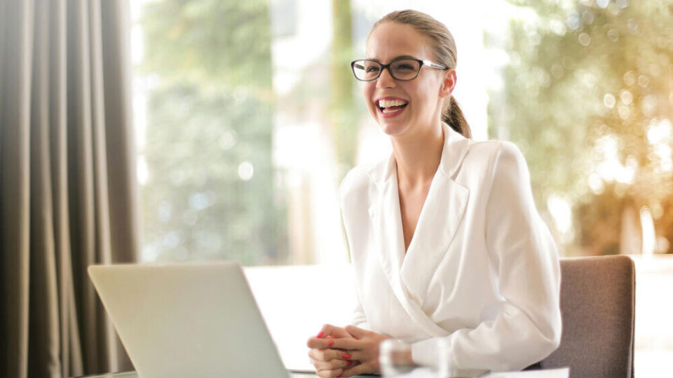 Lachende Frau mit Brille sitzt im Büro vor einem Laptop.