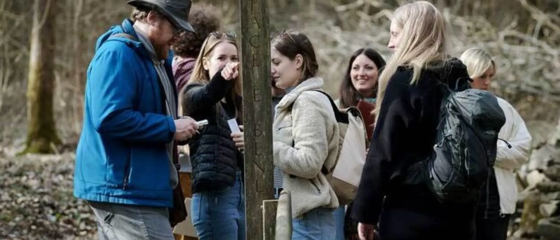 Gruppe von Menschen steht im Wald um einen hölzernen Wegweiser versammelt.