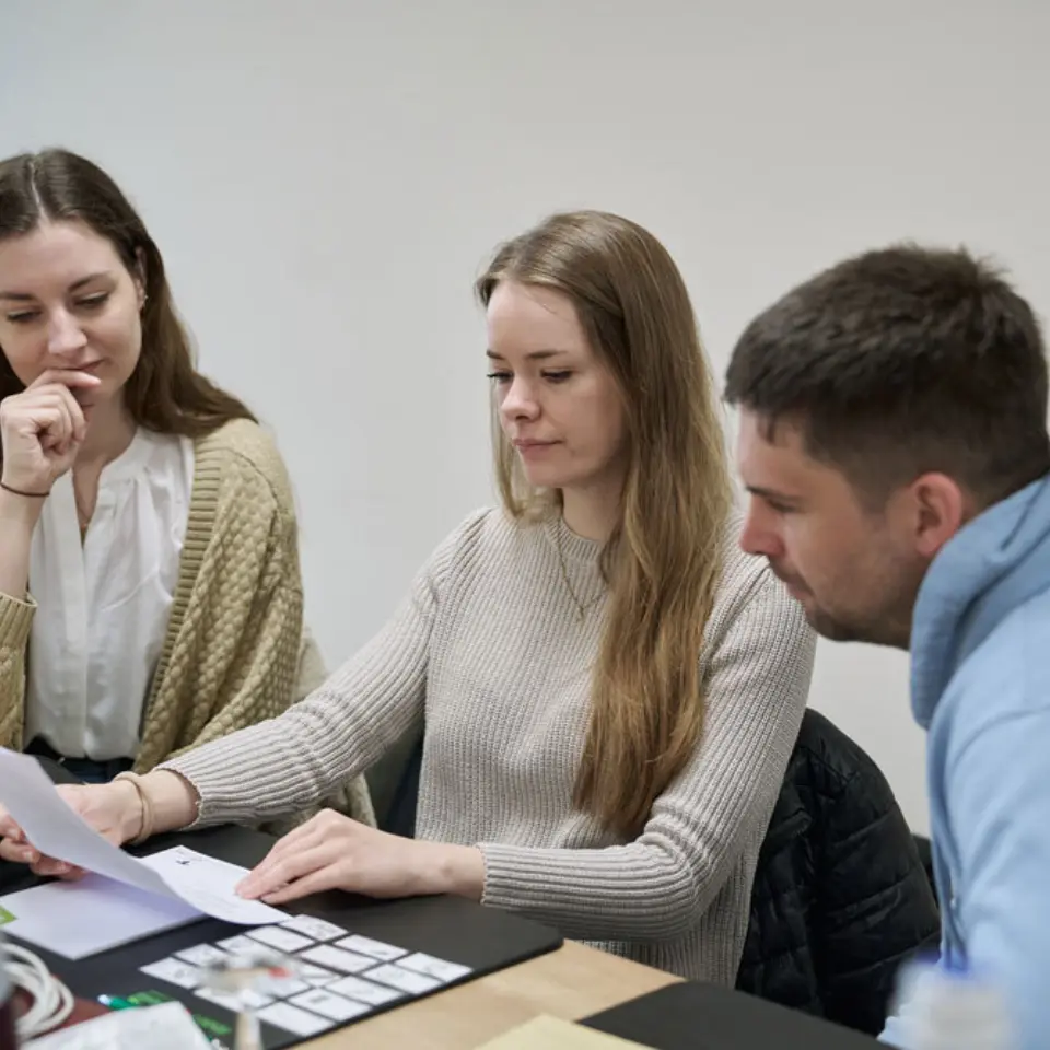 Drei junge Erwachsene sitzen am Tisch und betrachten gemeinsam ein Blatt Papier.