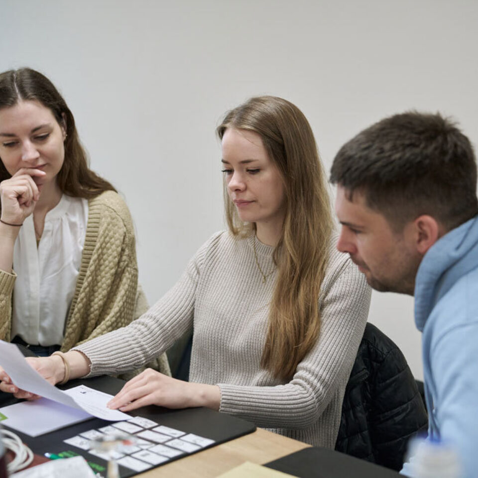 Drei junge Erwachsene sitzen am Tisch und betrachten gemeinsam ein Blatt Papier.