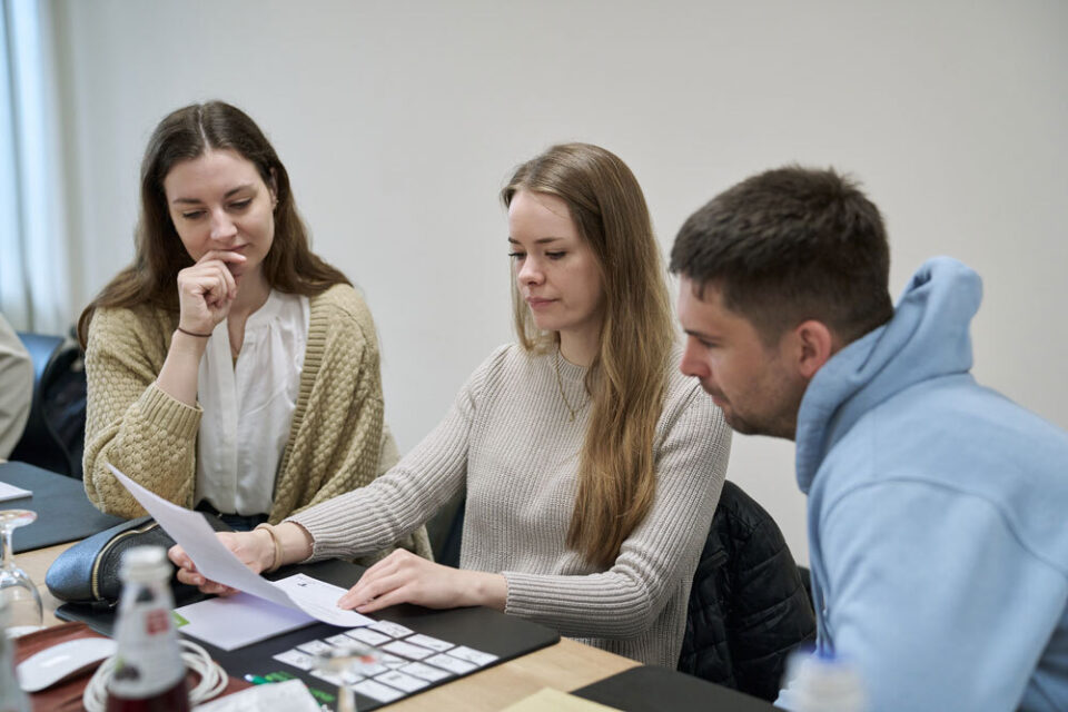 Drei junge Erwachsene sitzen am Tisch und betrachten gemeinsam ein Blatt Papier.