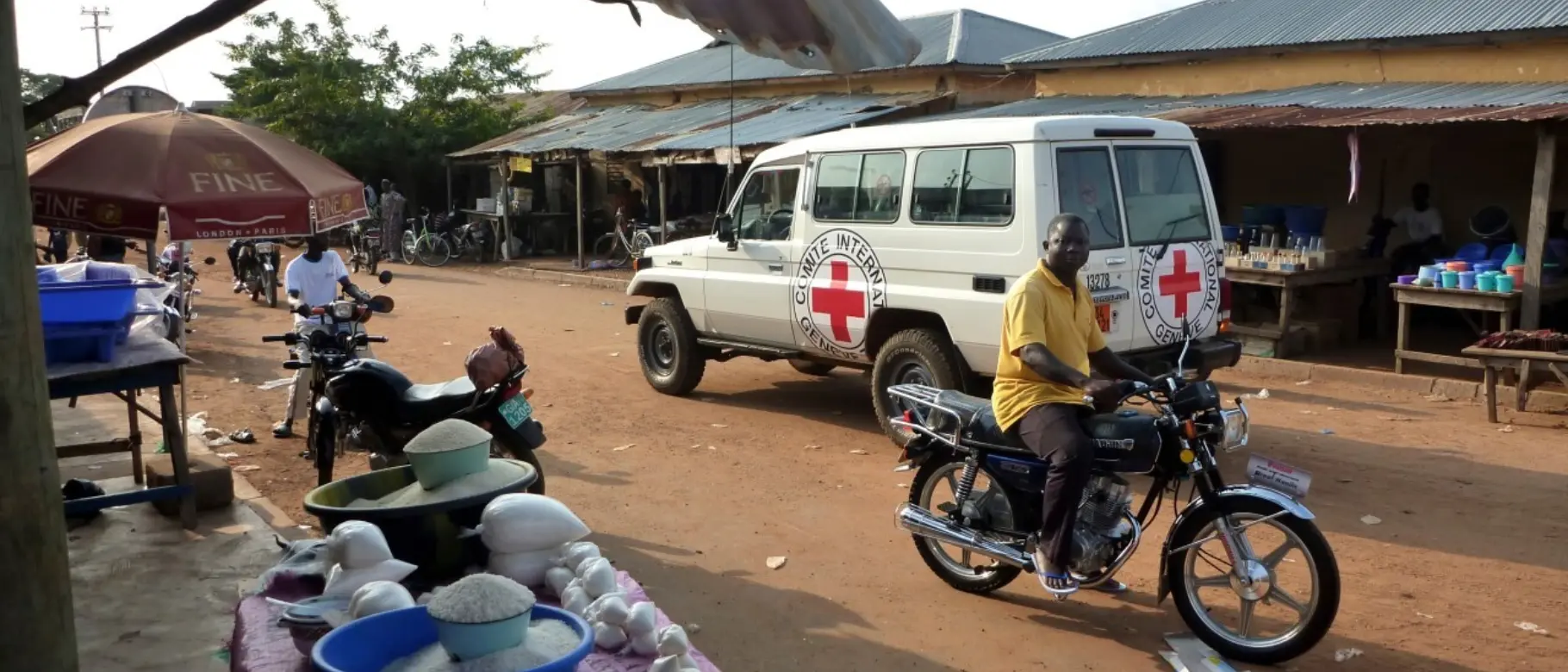 Straßenszene in Togo mit Fahrzeug des Internationalen Roten Kreuzes, Motorradfahrer und Verkaufsstand – als Bild für globale Zusammenhänge, Lieferketten und unternehmerische Verantwortung.