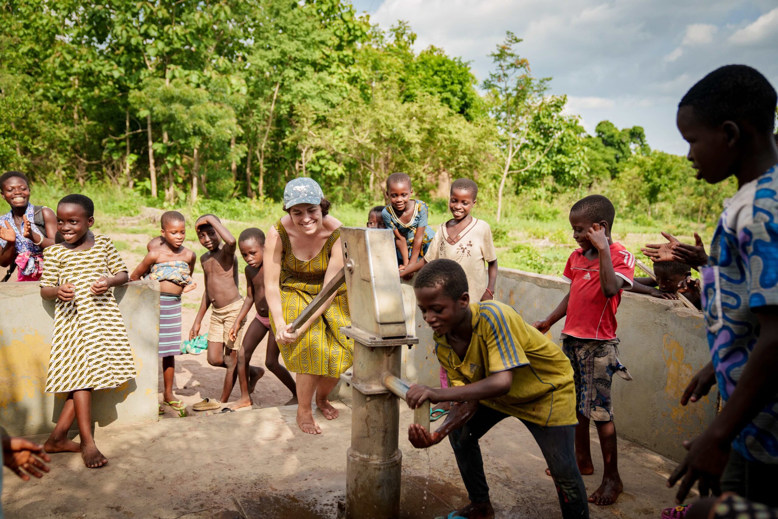 Kinder und eine Projektmitarbeiterin pumpen gemeinsam Wasser aus einem neu gebauten Brunnen im Rahmen von Project Togo – Freude und Zugang zu sauberem Trinkwasser.